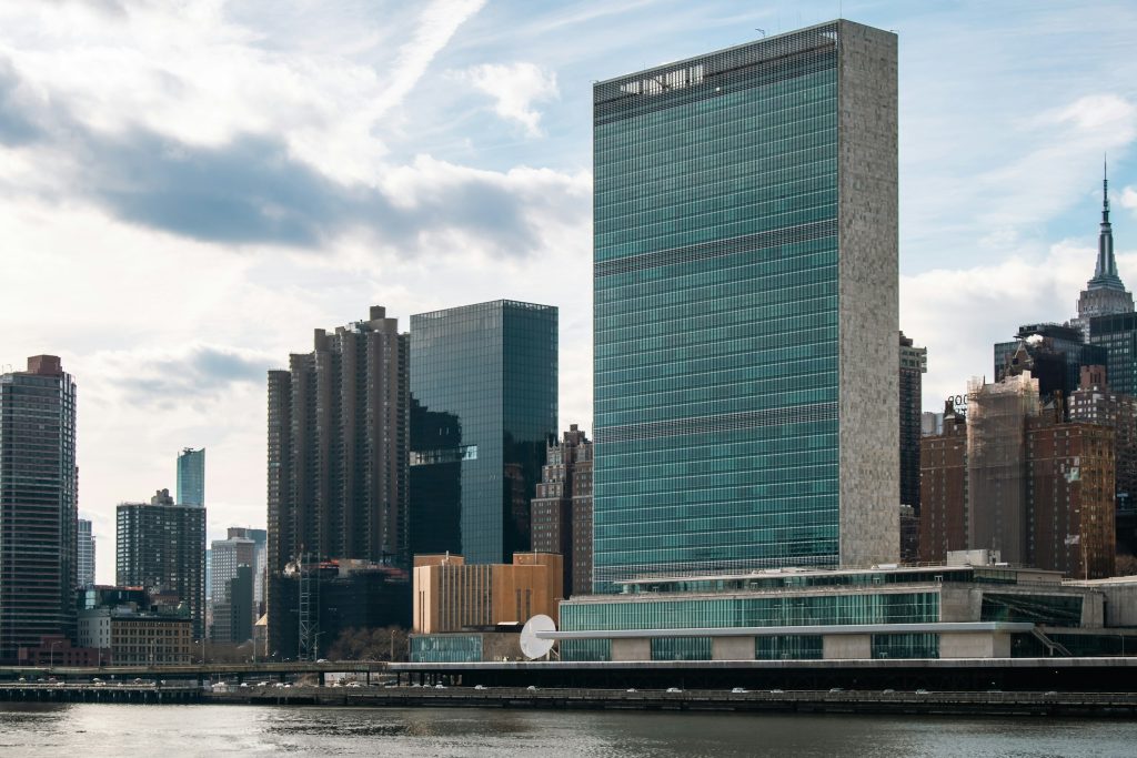 The United Nations Headquarters in New York City, seen from across the East River. The tall, glass-faced Secretariat Building stands prominently, embodying modernist architectural design. At its base lies the General Assembly Building, a horizontal structure where representatives from around the world convene. This iconic complex, situated in Midtown Manhattan, serves as a global hub for diplomacy and international policy. The waterfront view highlights the UN’s presence as both a political and architectural landmark in New York City.